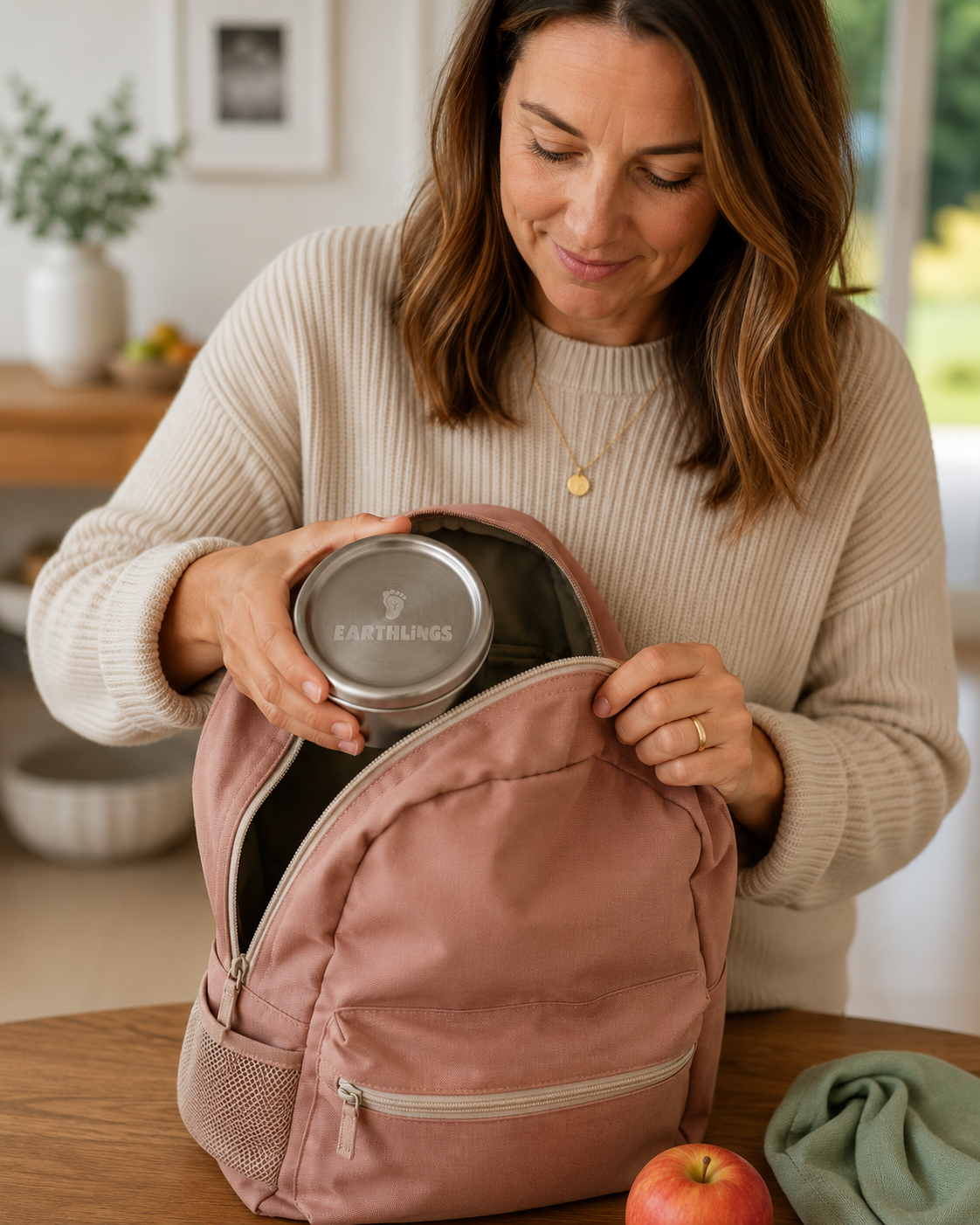 A woman in a beige ribbed sweater opens a dusty pink backpack on a wooden table, placing a stainless steel lunch container inside. An apple and a folded green cloth are visible nearby in a bright, modern kitchen setting.