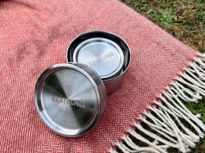 Two stainless steel Earthlings food storage containers, one open and one closed, resting on a red and white herringbone blanket outdoors on grass.