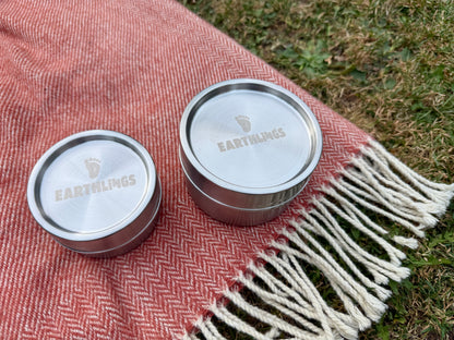 Two round metal tins with silver lids branded 'EARTHLINGS' resting on a red-and-white herringbone blanket with fringe, placed outdoors on grass.