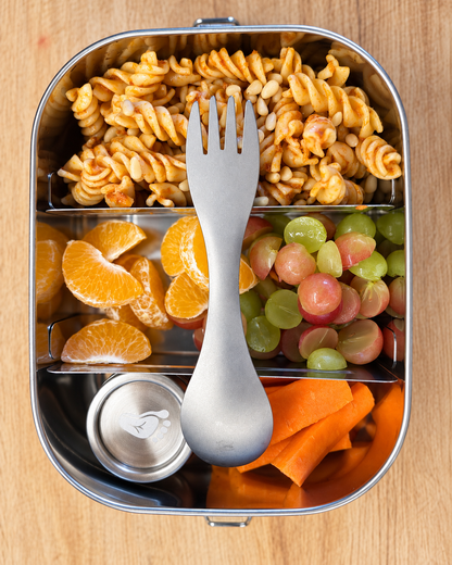 A stainless steel compartment lunch box on a wooden surface, filled with rotini pasta, mandarin slices, mixed grapes, carrot sticks, and a silver spork.